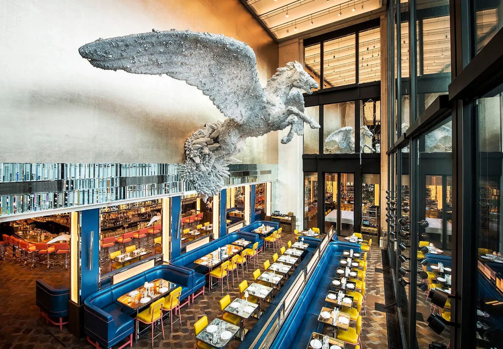 Damien Hirst's crystal-covered Pegasus sculpture suspended above the dining room at Brasserie of Light, blue booths and yellow chairs below