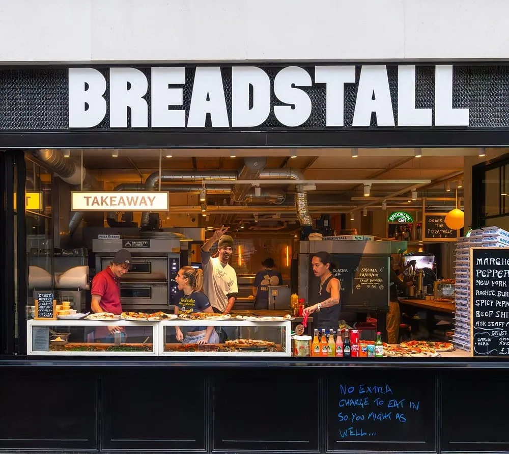 The Breadstall counter on Berwick Street in Soho, staff cutting large round pizzas behind a glass display with a menu board and stacked pizza boxes