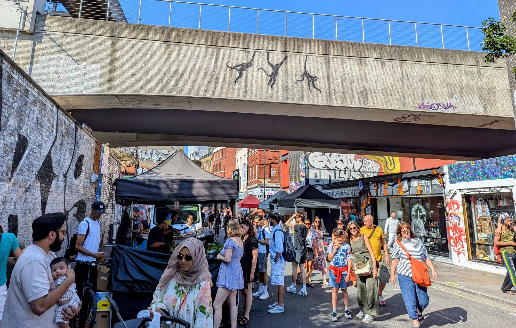 Market stalls filling Brick Lane beneath the railway bridge, with Banksy's three hanging monkeys stencilled on the concrete above