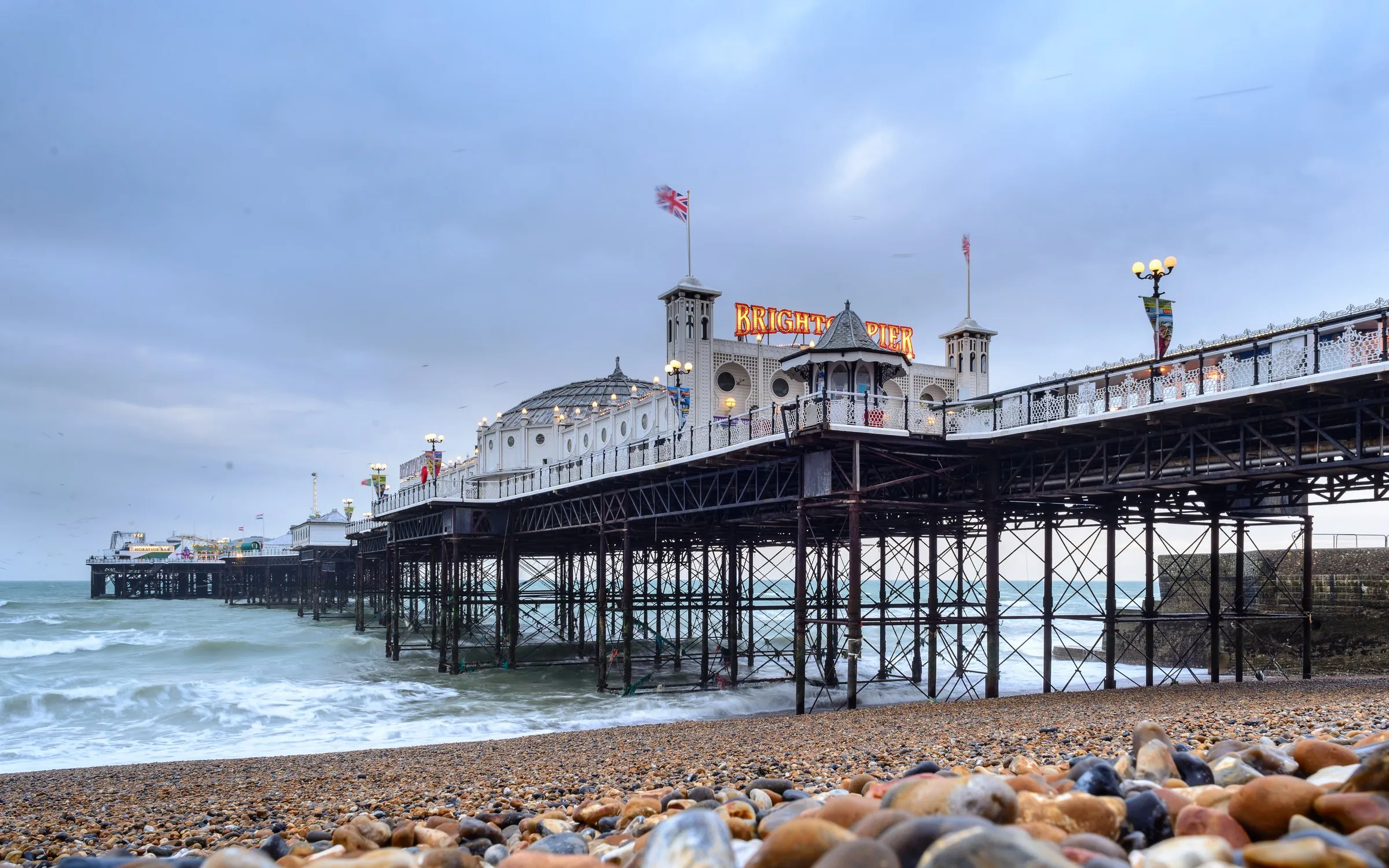 Brighton Pier at dusk, its lit sign and arcade building on stilts over the pebble beach