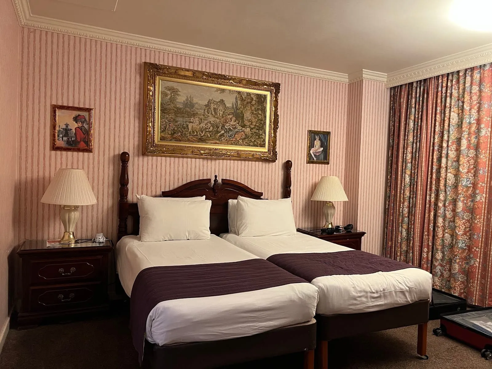 A twin room at the Britannia International Hotel with two single beds pushed together under a carved wooden headboard, a gilt-framed tapestry, pink striped wallpaper, cream table lamps and floral curtains