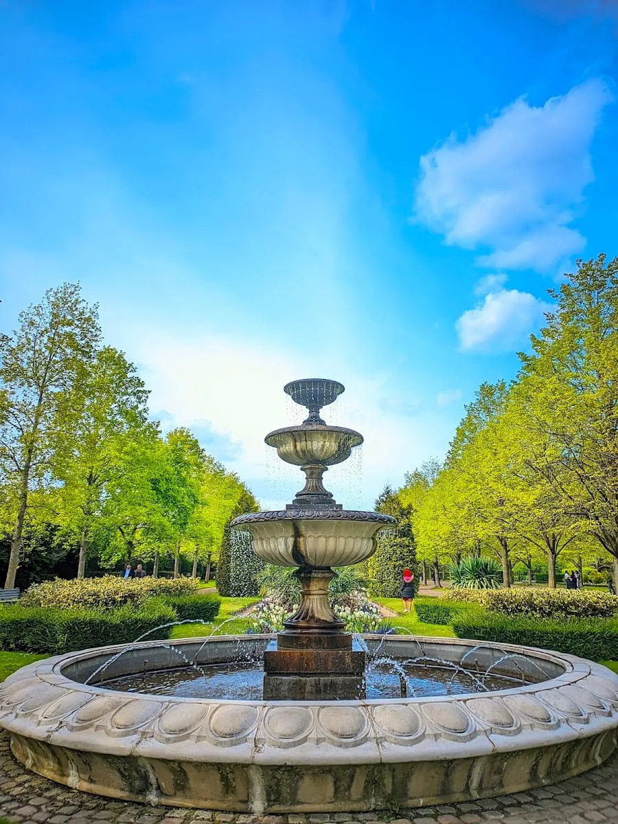 A three-tiered stone fountain with water jets, framed by trees along the Avenue Gardens