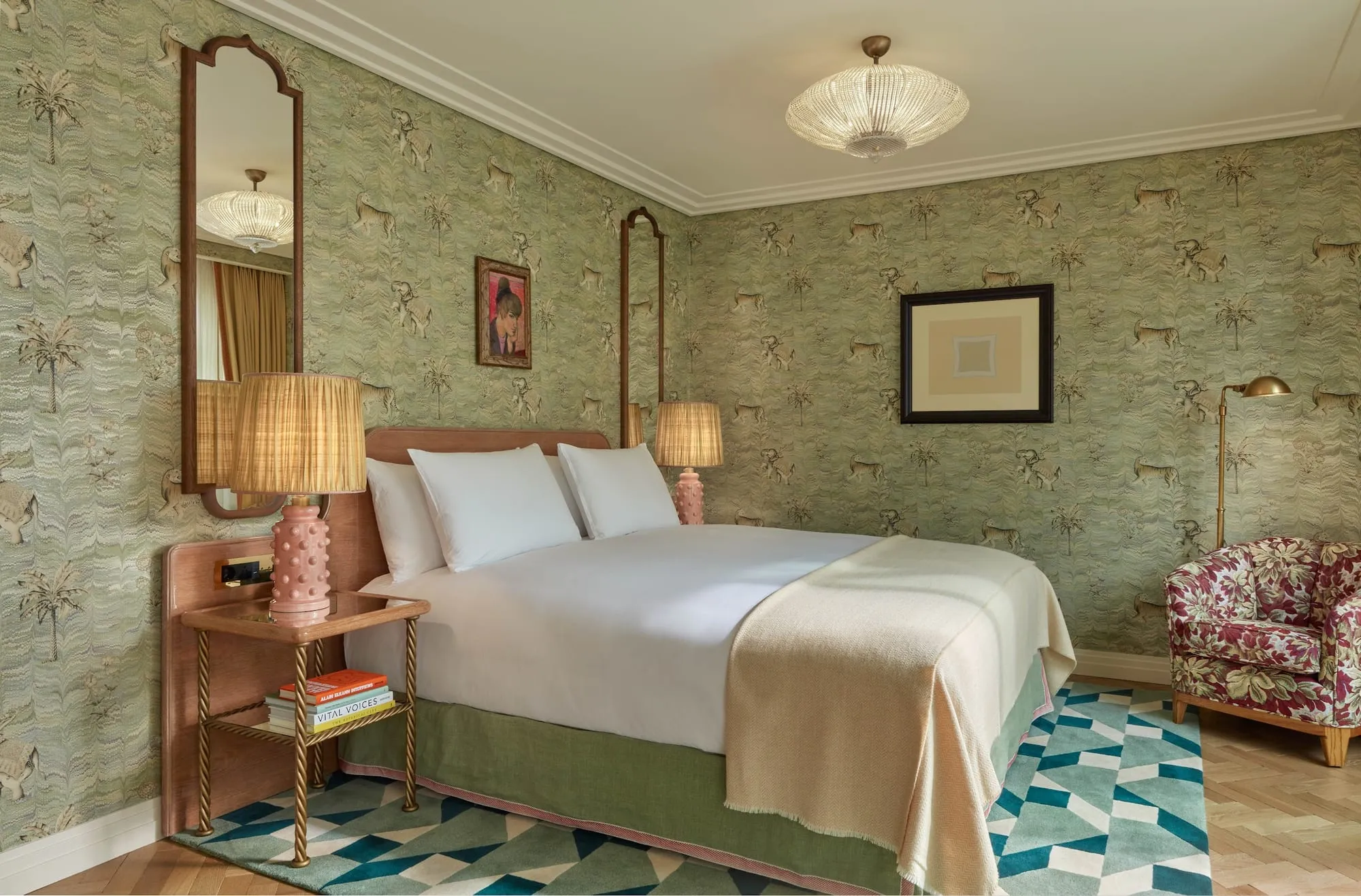 A Broadwick Soho bedroom with green wallpaper patterned with tigers, elephants and palm trees, an ornate arched mirror, pink table lamps and a floral armchair