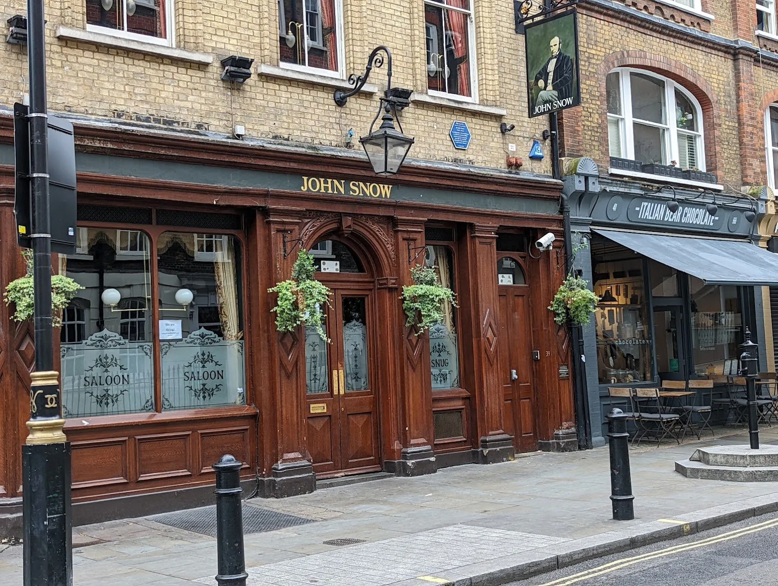 The John Snow pub on Broadwick Street: a dark wood Victorian frontage with etched Saloon and Snug windows, hanging baskets, a lantern, and a pub sign painted with Snow's portrait
