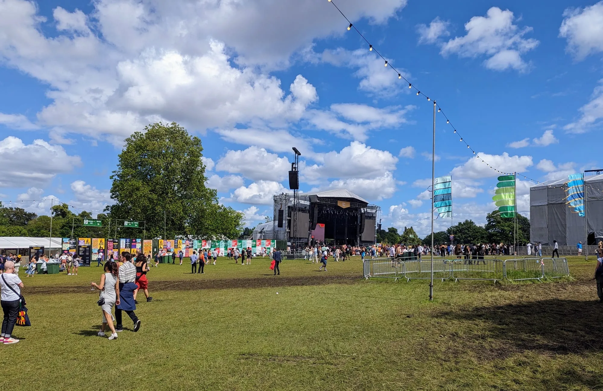 Cross the Tracks in Brockwell Park: people crossing the grass towards a covered main stage, with bunting, flags and a row of colourful stalls under a blue sky