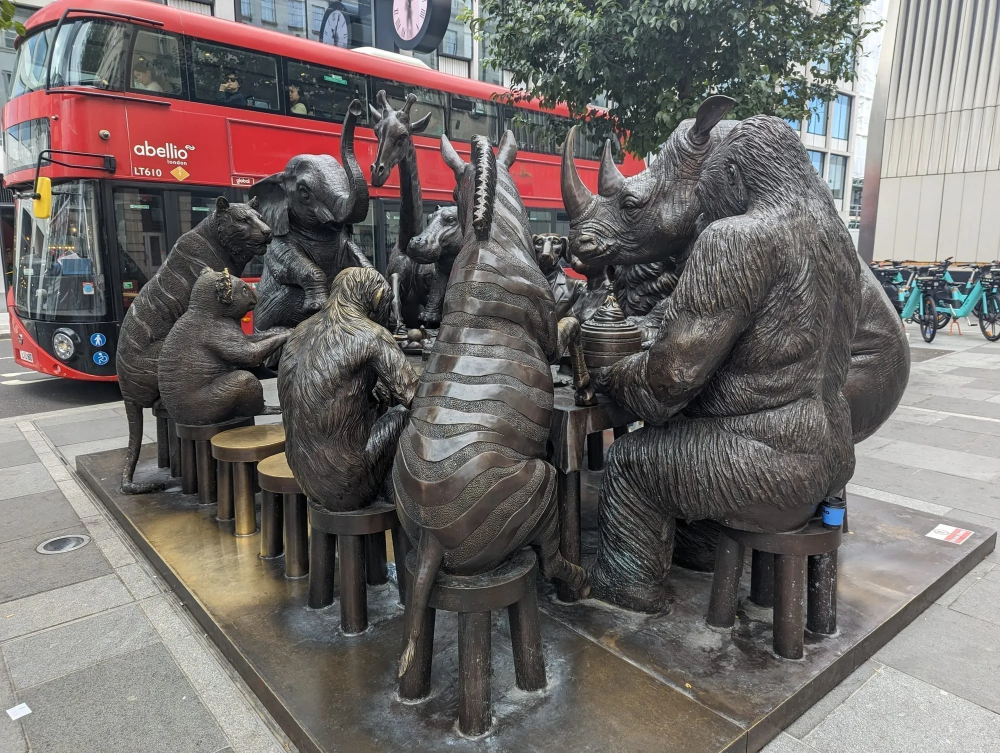 A bronze sculpture of animals seated around a dining table on the plaza outside Paddington station, with a red London bus passing behind