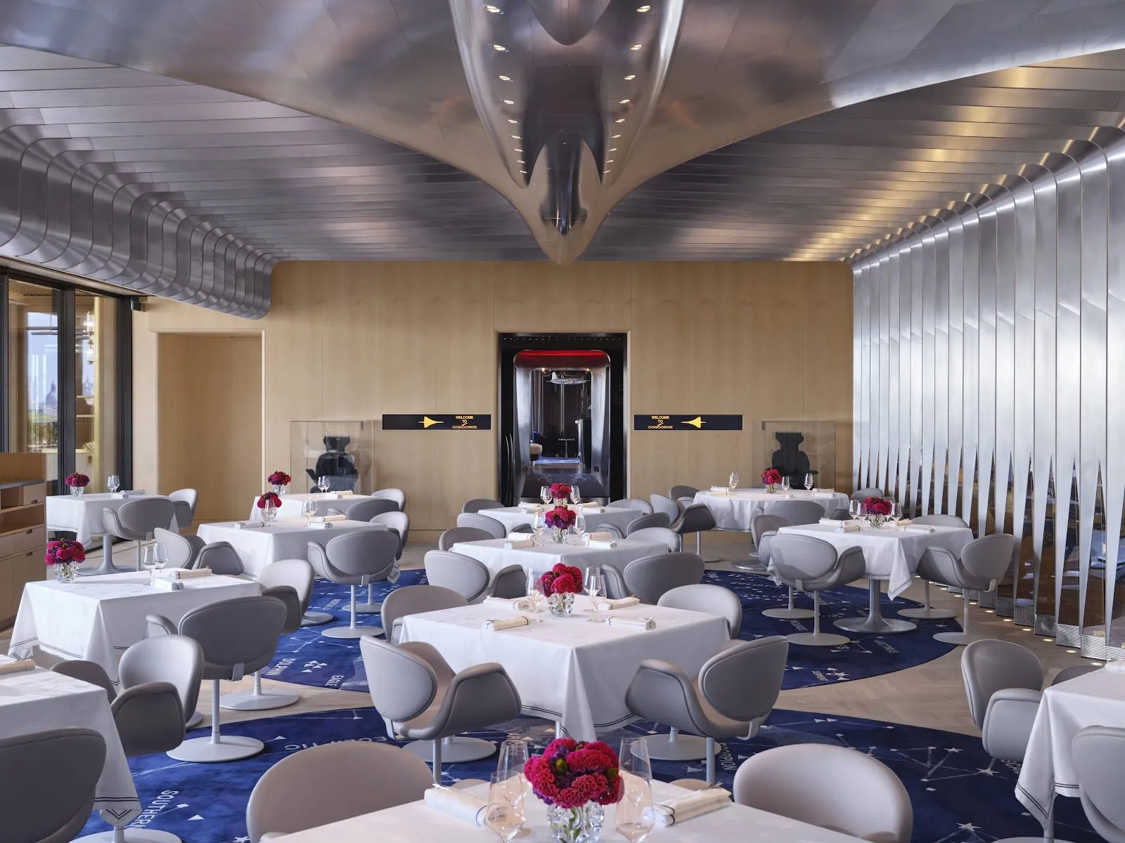 The Brooklands dining room, with a polished aluminium Concorde fuselage running across the ceiling, white-clothed tables and grey tulip chairs on a navy star-chart carpet, and a doorway marked "Welcome to Concorde" at the far end