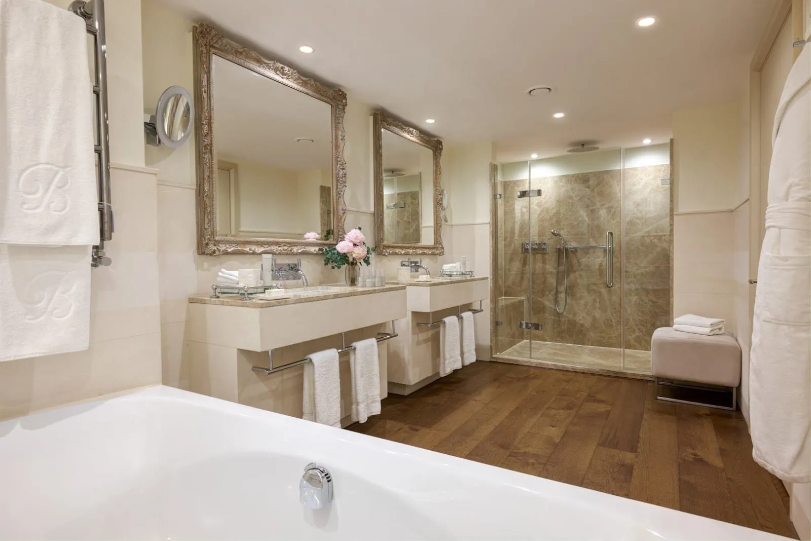 A Brown's Hotel bathroom with two stone vanities under carved gilt mirrors, a wooden floor, a walk-in shower lined in brown marble and a bath in the foreground