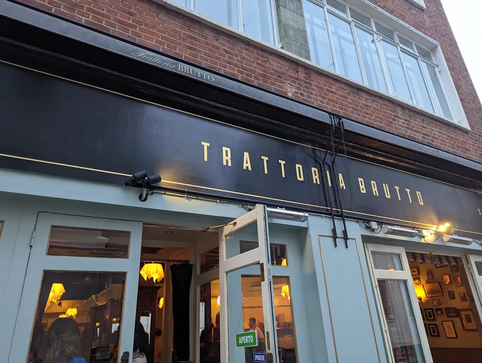 The gold-lettered Trattoria Brutto sign above pale green shopfront doors, with an Aperto sign hanging