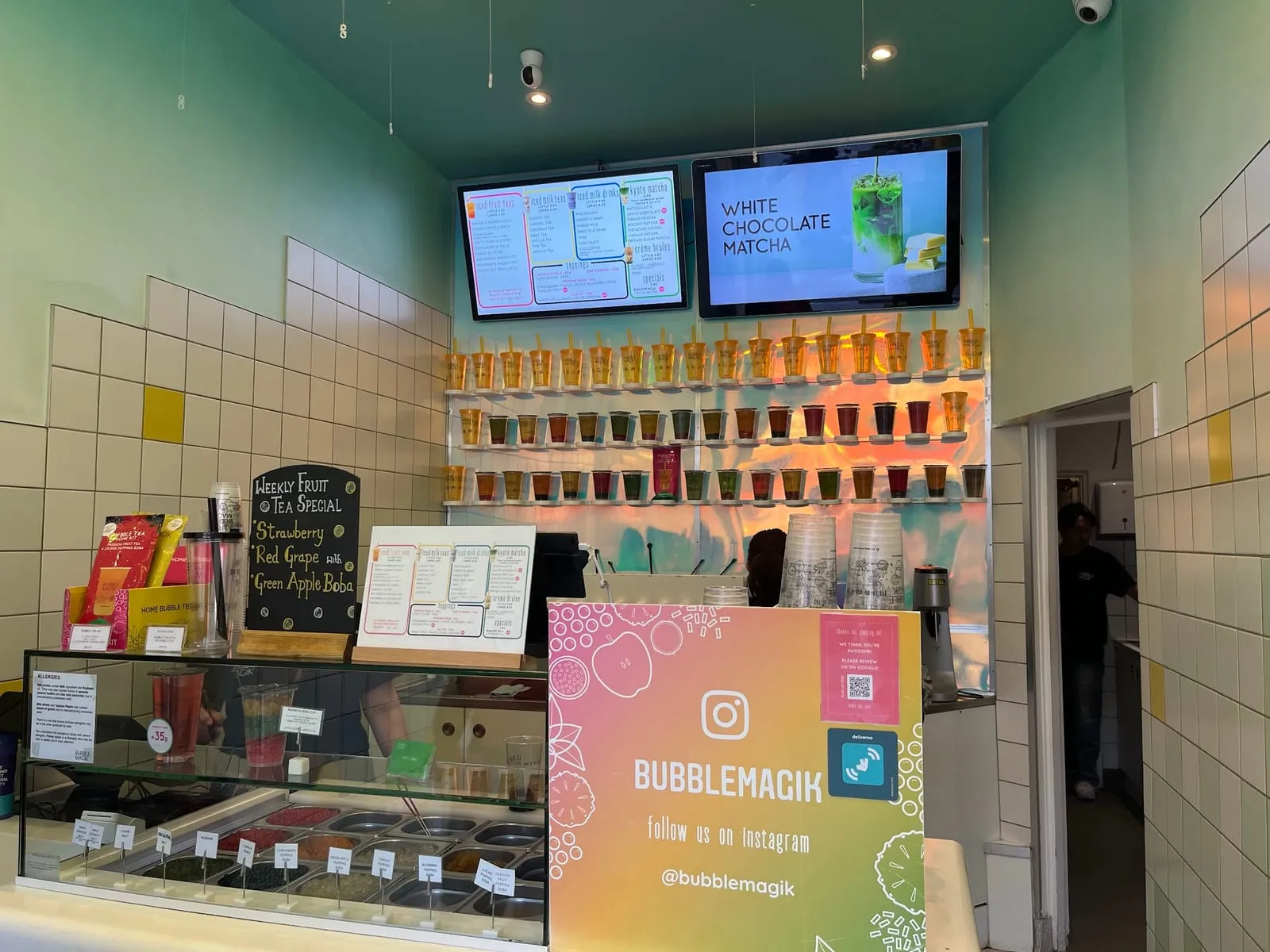 Inside Bubble Magik: a chilled display of fruit toppings at the front of the counter, menu screens above and a wall of sealed cups behind