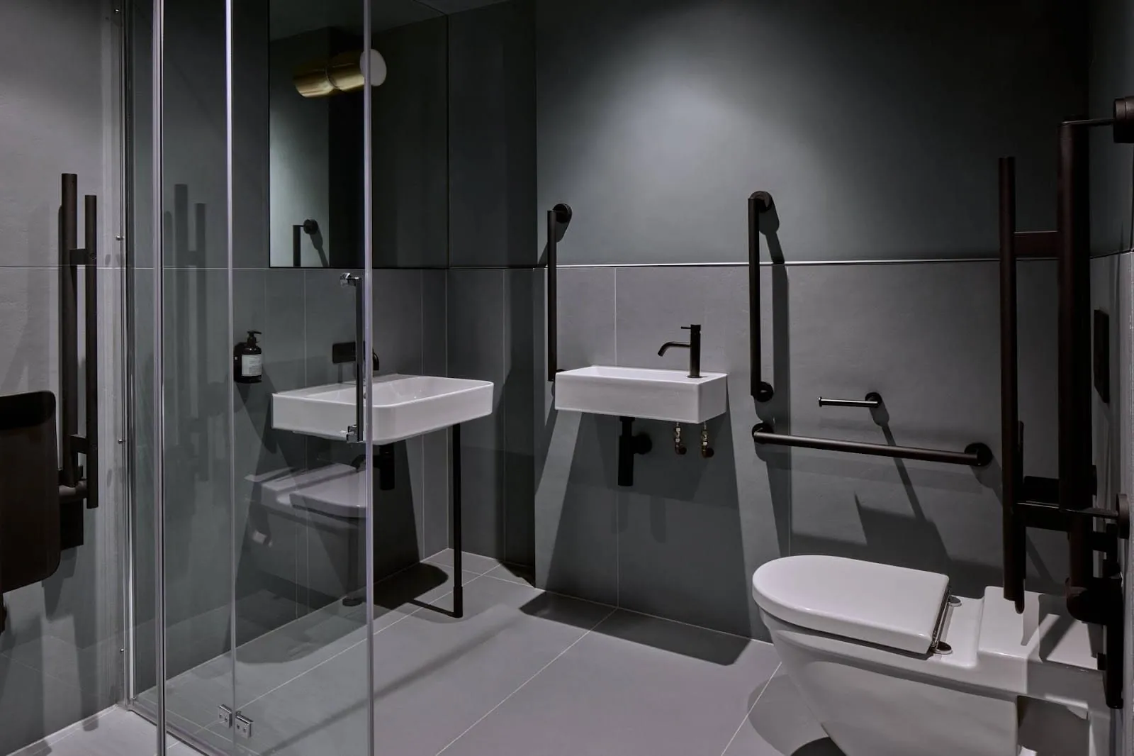 An accessible bathroom at Buckle Street Studios in charcoal grey, with black grab rails, a fold-down shower seat behind glass, two white basins and a wall-hung toilet