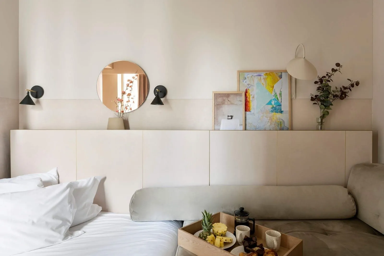 A room at Buckle Street Studios with a white bed and a velvet daybed sharing one long pale headboard, black wall lights, a round mirror and a breakfast tray with pineapple and a cafetière