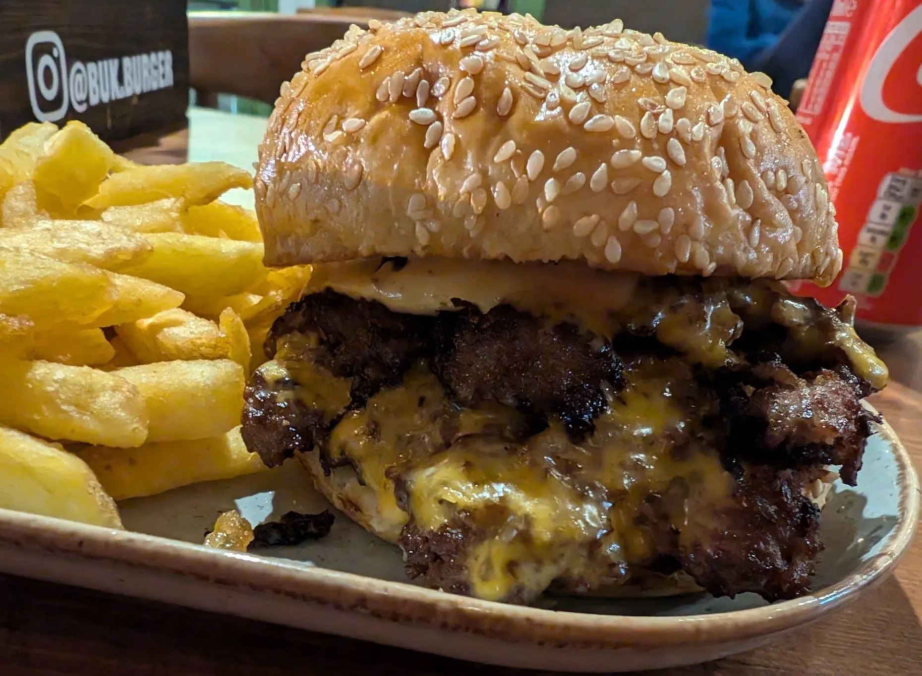 A double cheeseburger with melted cheese in a sesame bun, beside thick-cut chips and a bottle of Coca-Cola, with a sign for Buk Burger's Instagram handle on the wall behind