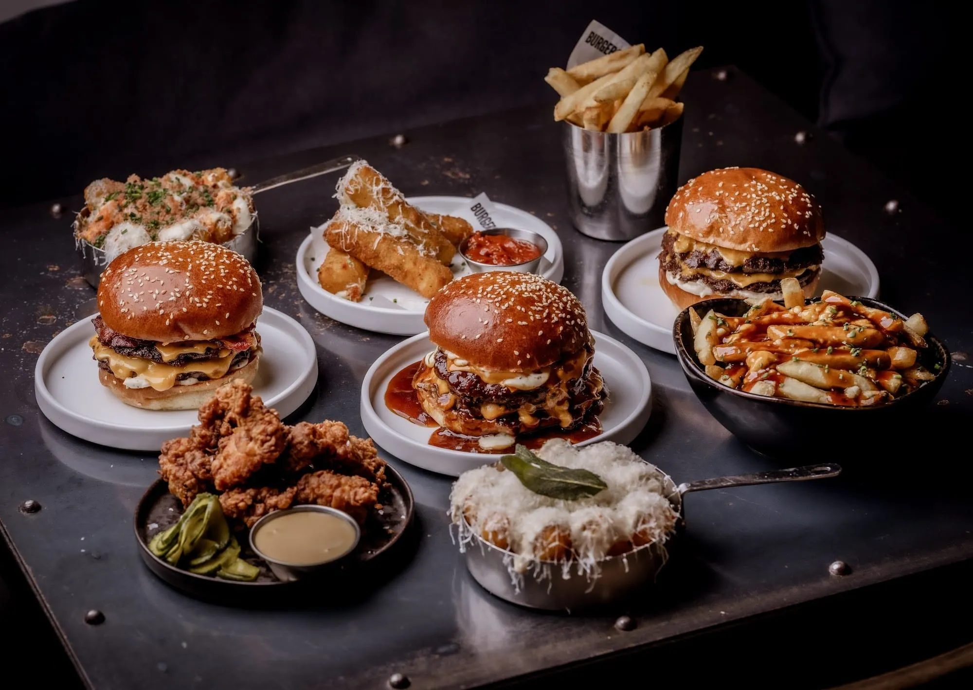 A spread of burgers, fried chicken tenders, mozzarella sticks and a bowl of loaded fries laid out on a dark metal table