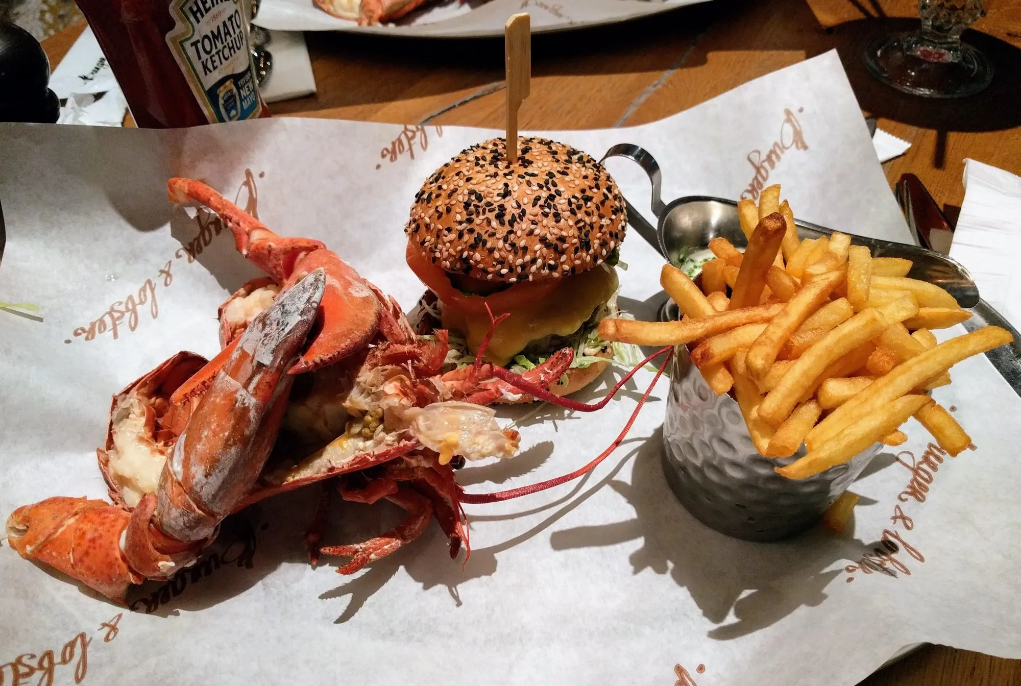 Half a lobster, a sesame-seed cheeseburger on a skewer and a metal cup of fries, laid out on Burger & Lobster branded greaseproof paper on a wooden table
