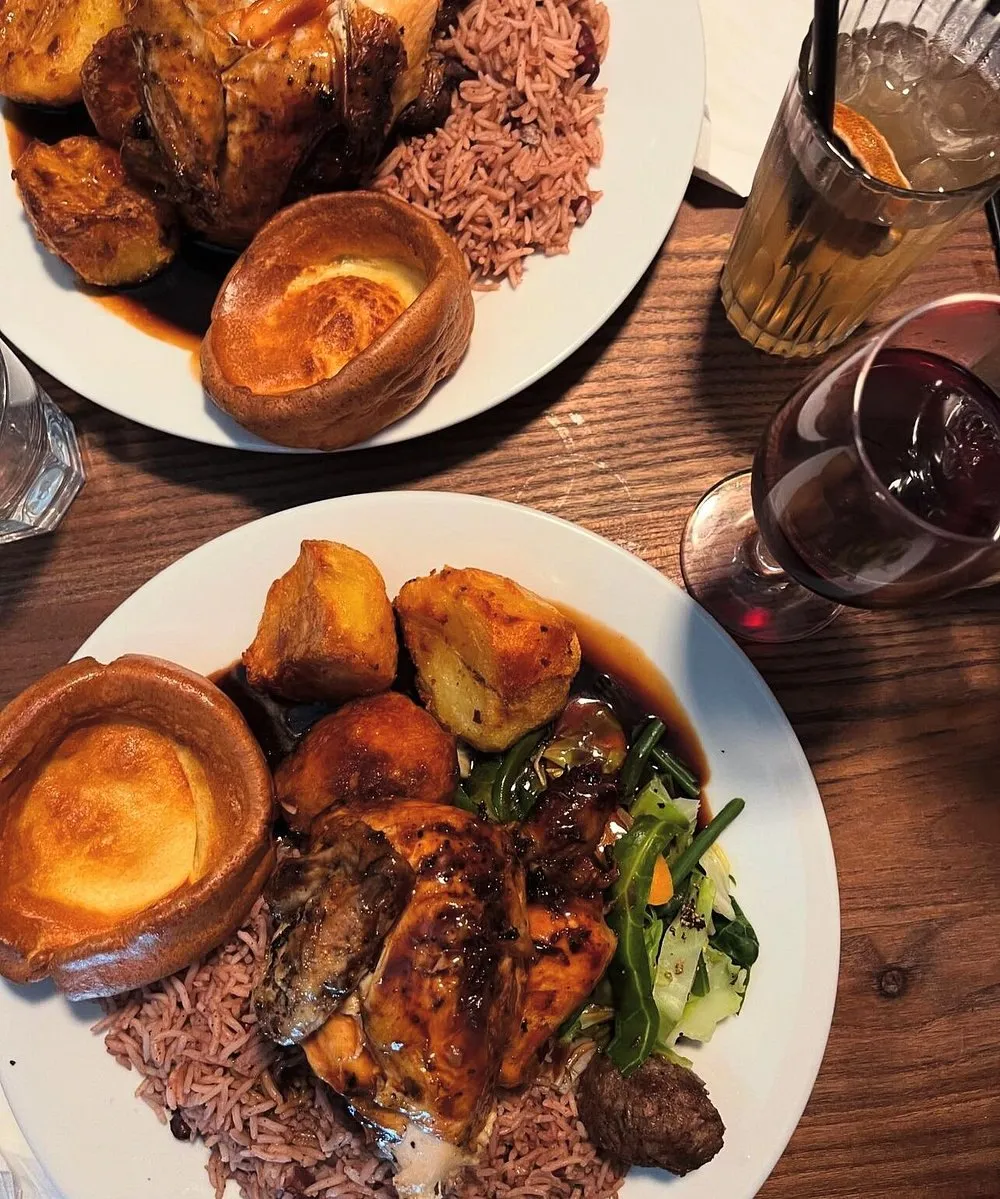 Two plates of Caribbean Sunday roast - glazed jerk chicken over rice and peas with roast potatoes, greens, gravy and a Yorkshire pudding - with a glass of red wine alongside