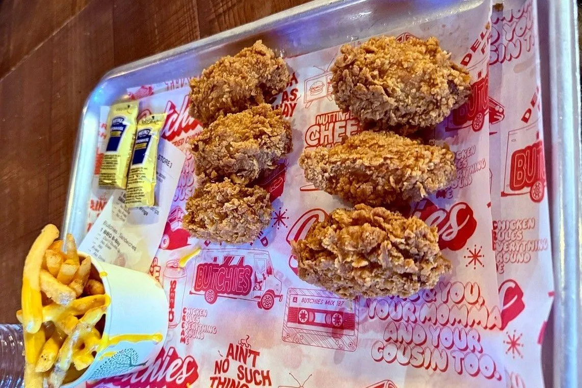 Buttermilk-fried chicken tenders on a metal tray lined with branded paper, with cheese-covered fries alongside
