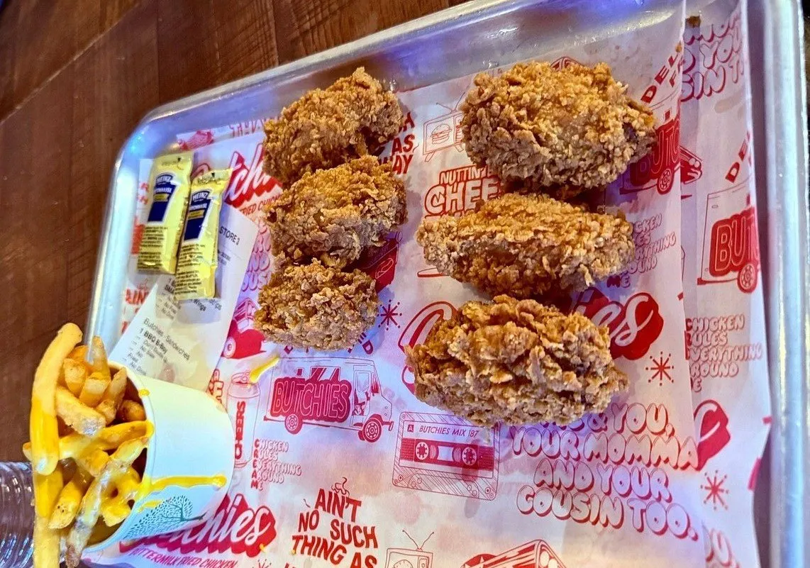 Six buttermilk-fried chicken tenders on a metal tray lined with Butchies branded paper, with a cup of cheese fries and mayonnaise sachets