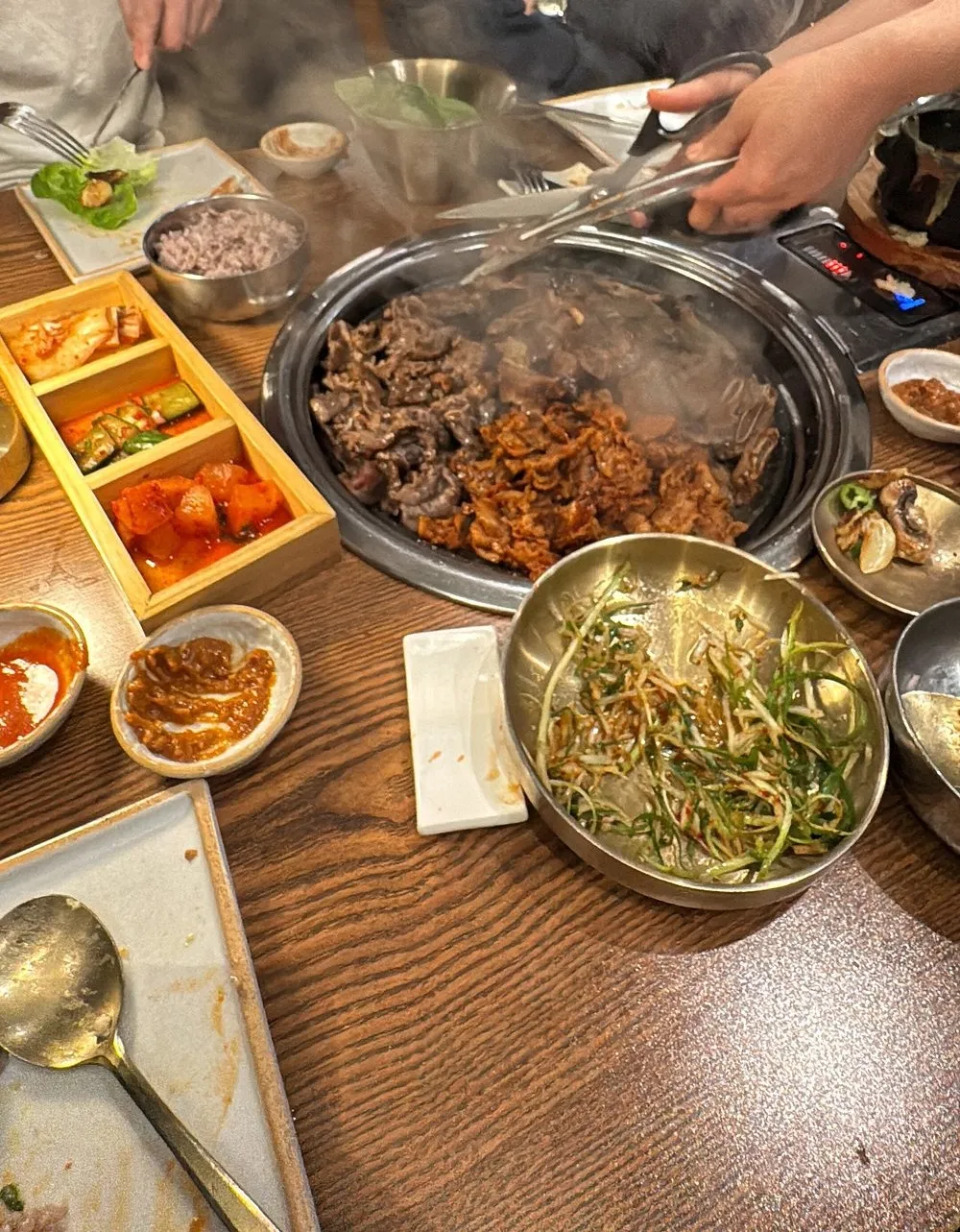 Marinated and plain beef cooking on a domed table grill at Cah Chi, with a wooden box of three kimchis, a bowl of purple rice, dipping sauces and a bowl of spring onion salad