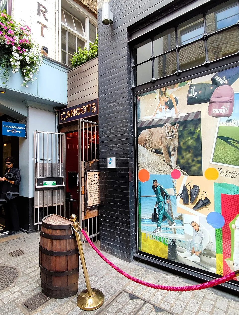 The entrance to Cahoots in Kingly Court, with CAHOOTS lit over a doorway behind an iron gate, an Underground sign beside it, a barrel and a rope barrier on the cobbles