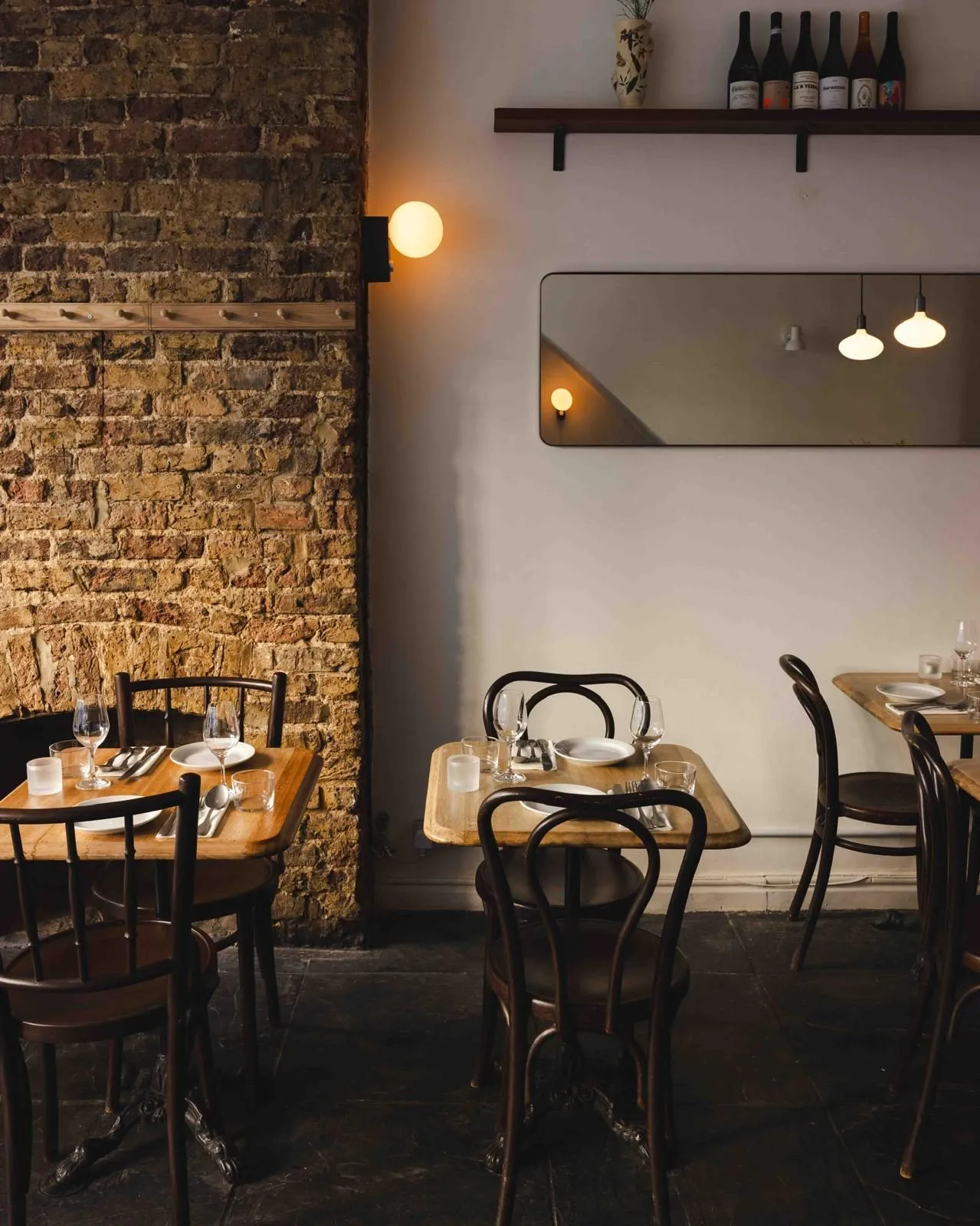 Bentwood chairs and small wooden tables against an exposed brick chimney breast at Cálong, with a globe wall light and a shelf of natural wine bottles