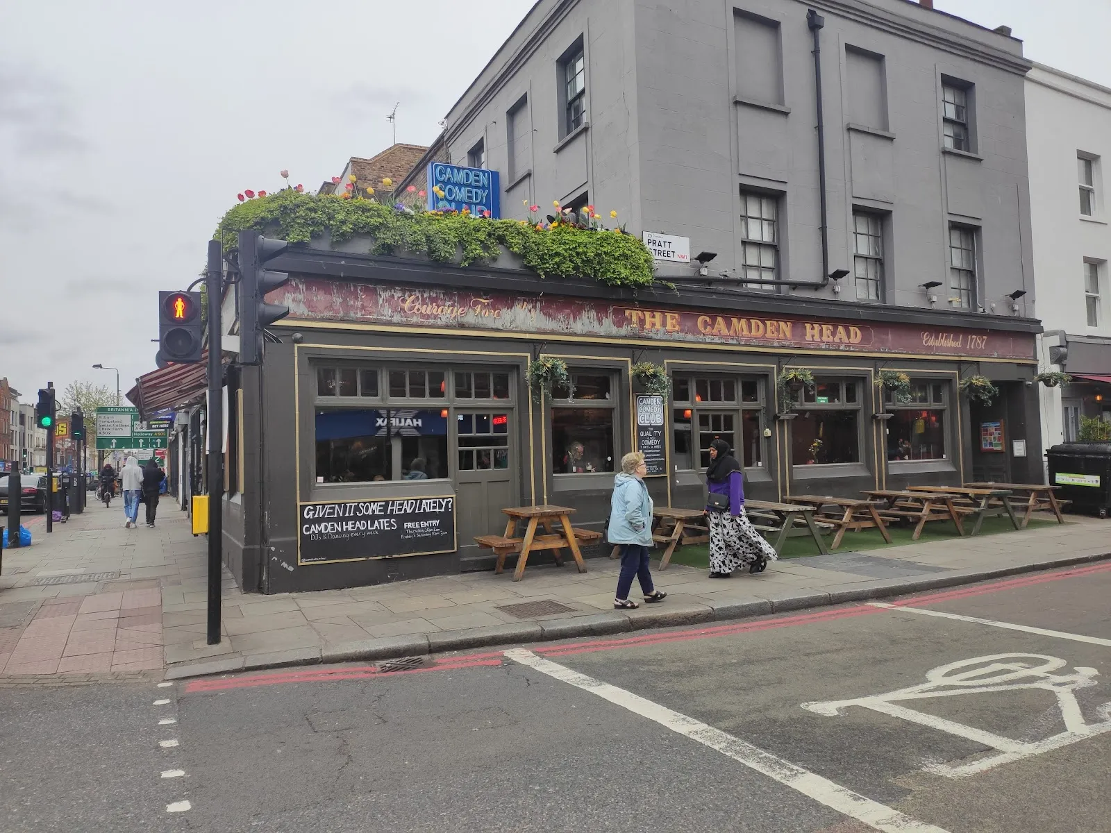 The corner exterior of The Camden Head pub, established 1787, with a blue "Camden Comedy Club" sign on the roofline above hanging flower baskets