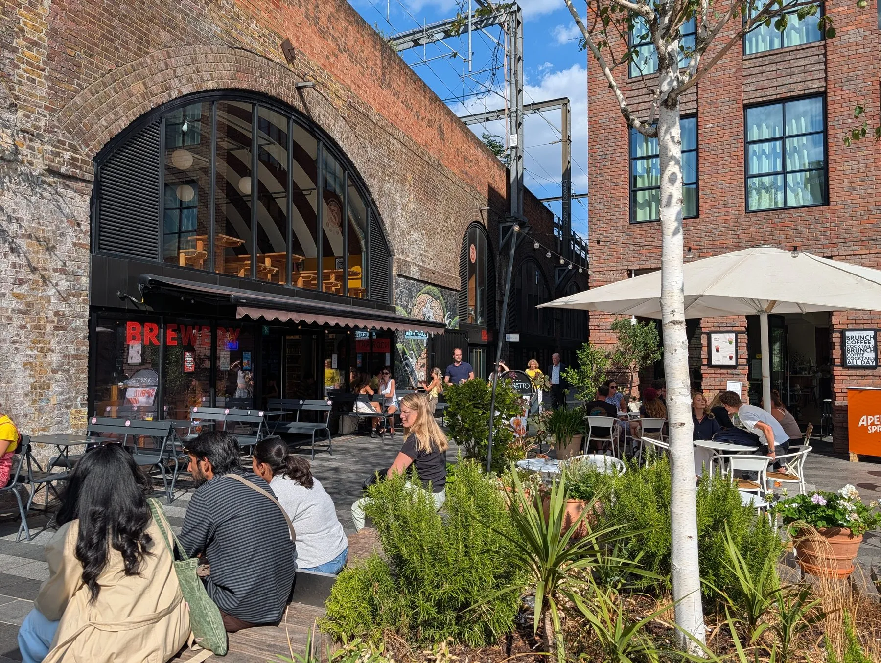 Outdoor tables under brick railway arches at Hawley Wharf in Camden, with a brewery tap room and beer hall behind and overhead railway wires above