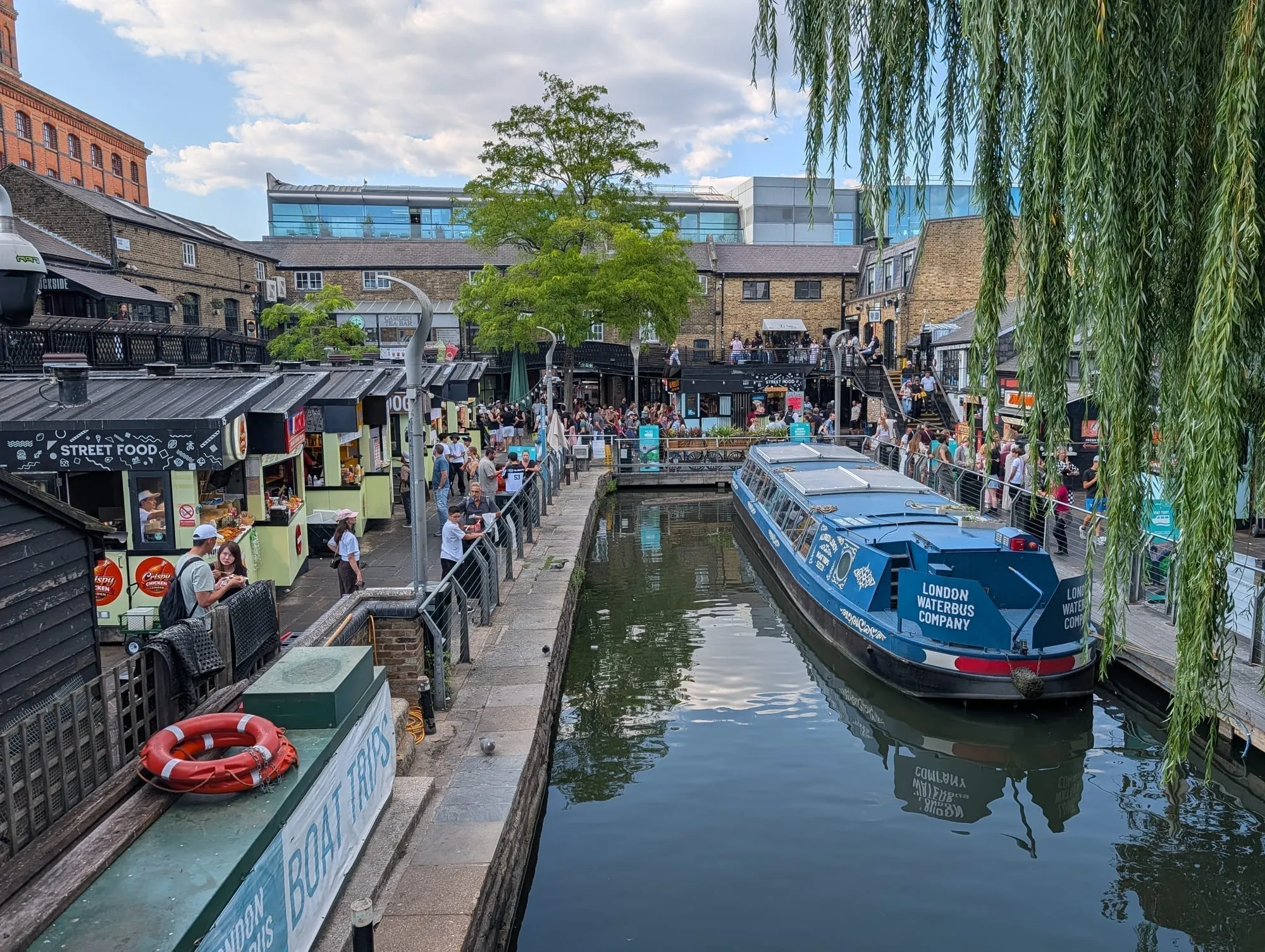 A narrowboat moored at Camden Lock beneath a willow tree, with street food counters along the far bank