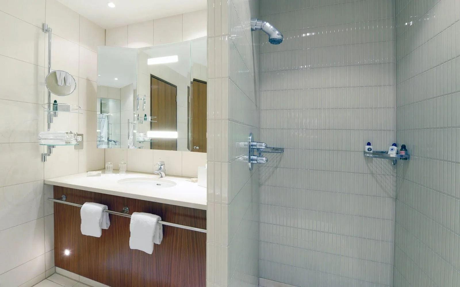 A bathroom at Canary Riverside Plaza with a walk-in shower tiled in small white tiles, a white vanity over a walnut cabinet, and a three-panel mirror