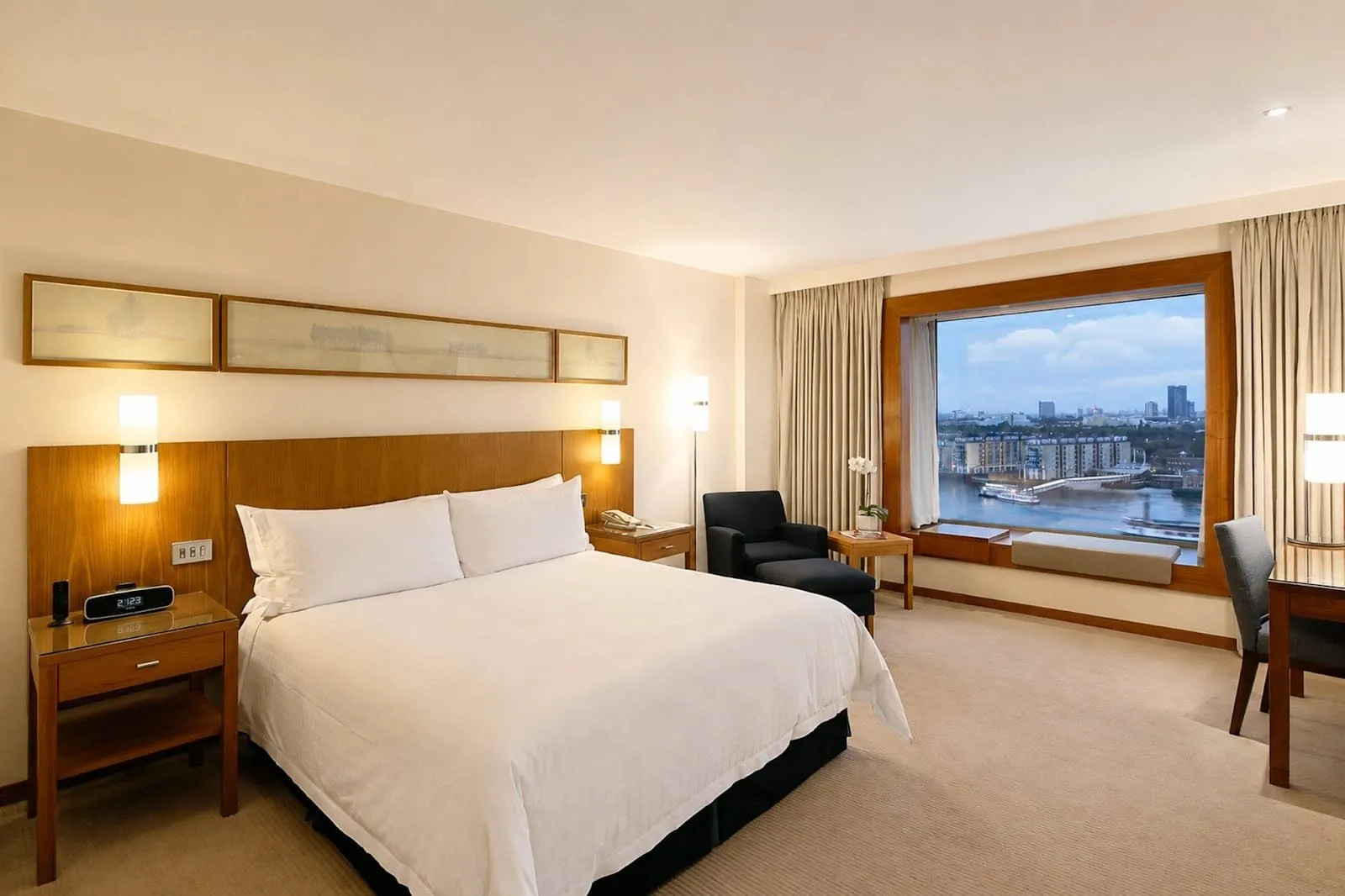 A king room at Canary Riverside Plaza with a walnut headboard, a black armchair and footstool, and a wide window seat looking across the Thames to riverside flats and moored boats