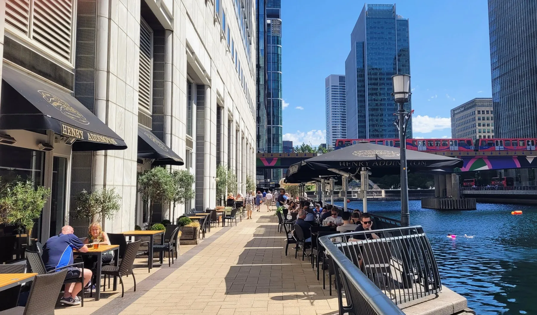 Tables along the dockside at Canary Wharf outside the Henry Addington, with a DLR train crossing the water and towers behind