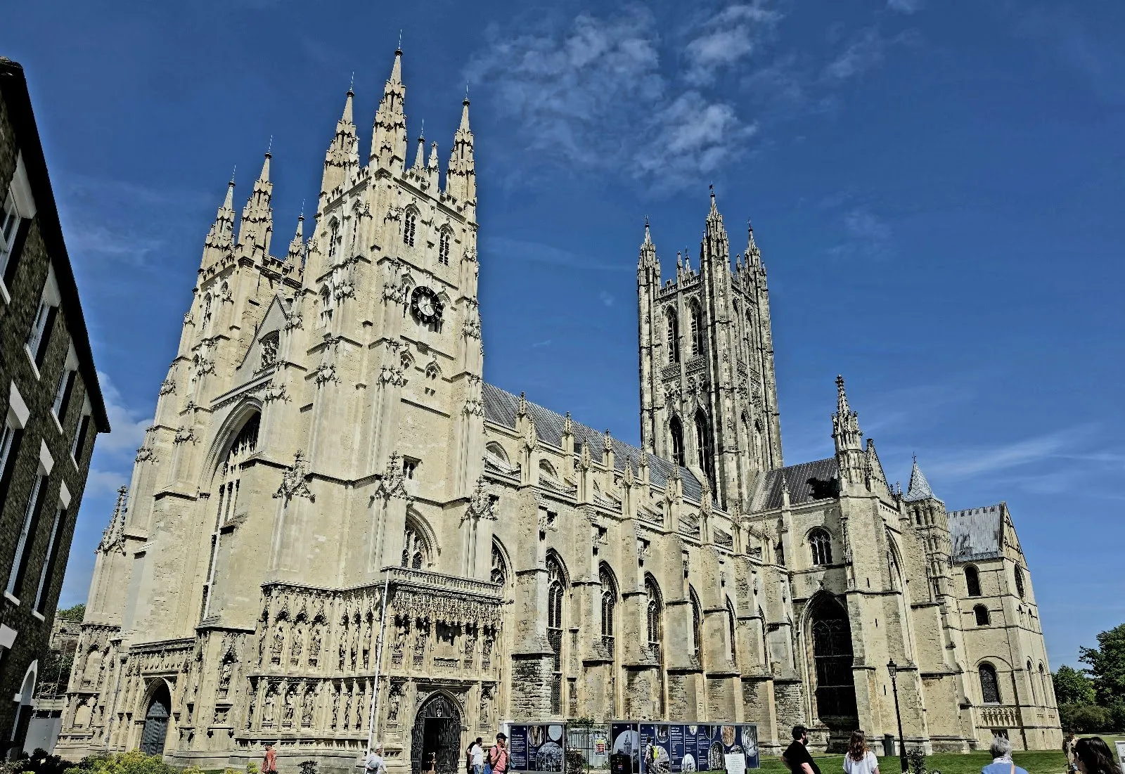 Canterbury Cathedral's west towers and central Bell Harry tower under a clear blue sky, with visitors on the lawn in front