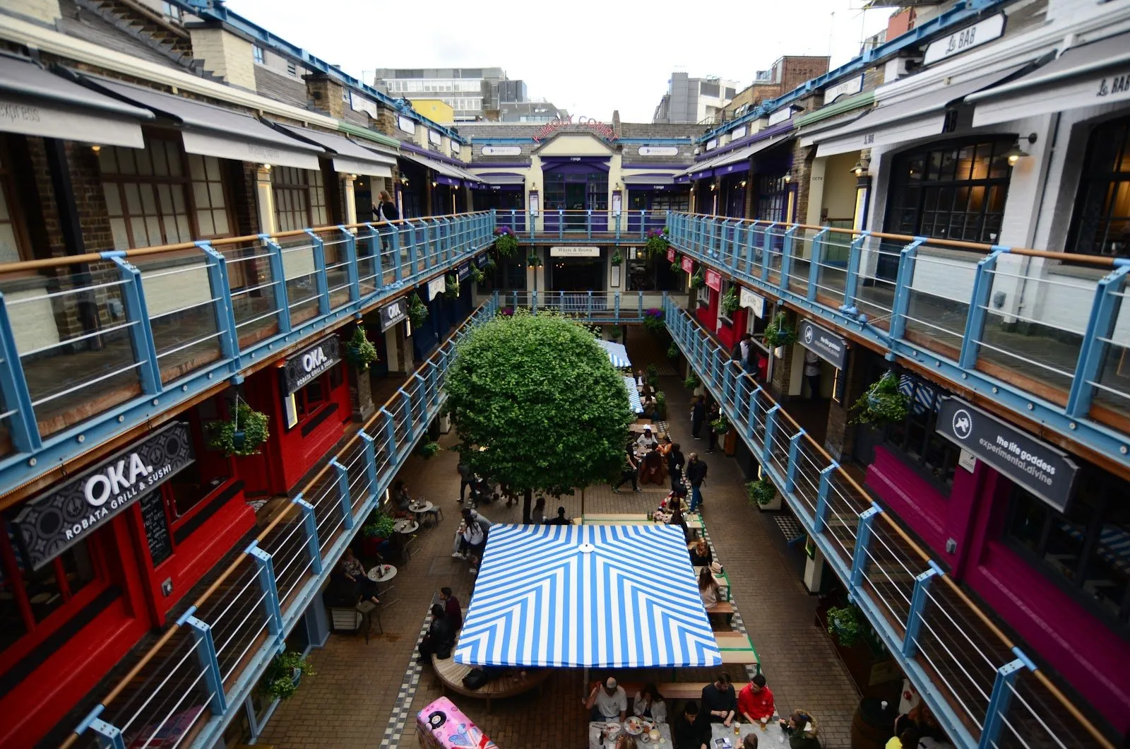 Kingly Court from an upper walkway: three floors of red, pink and grey restaurant fronts behind blue railings, around a courtyard with a round-clipped tree, tables and a blue-and-white striped canopy