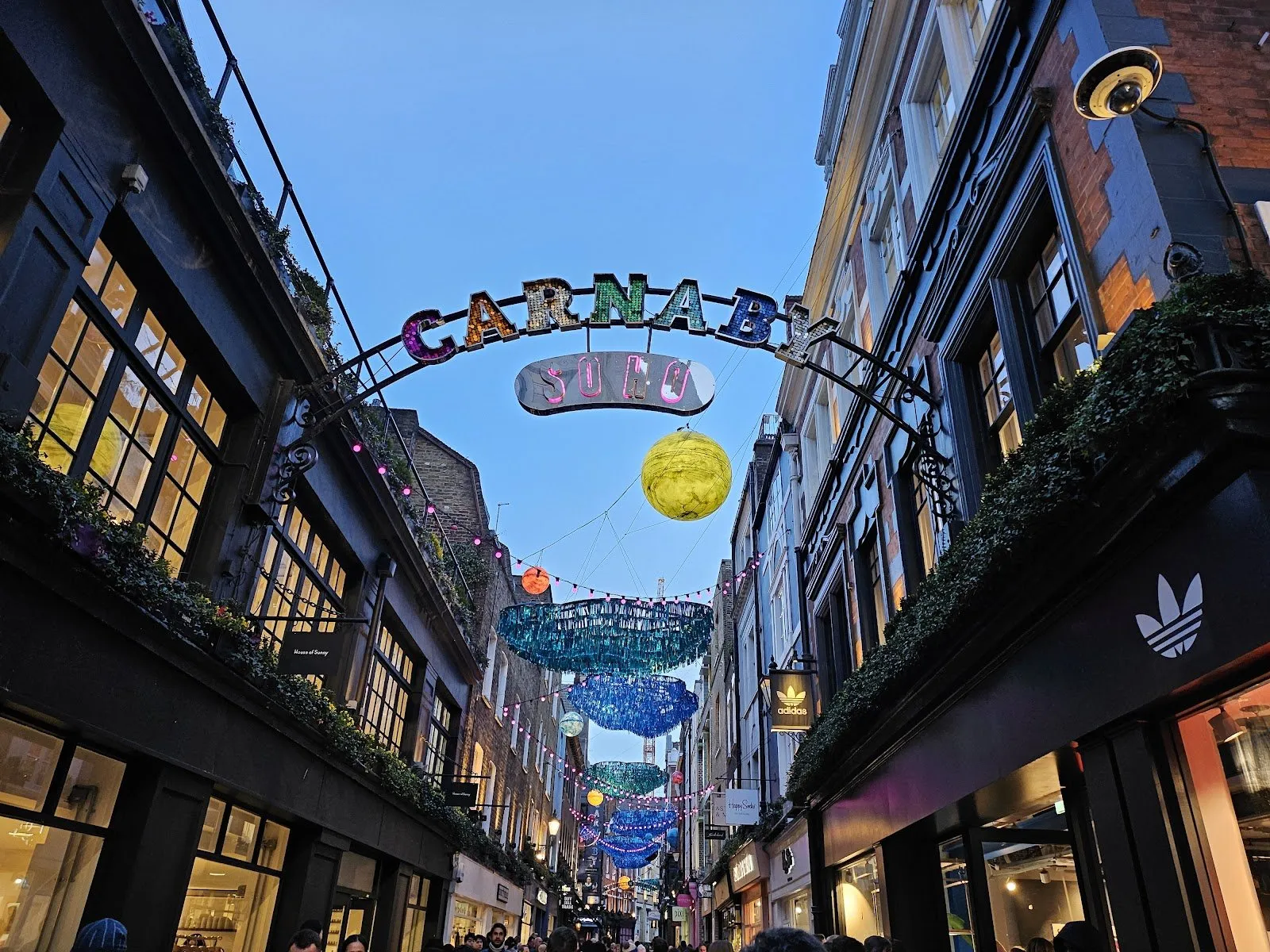 The Carnaby Soho sign arching over Carnaby Street at dusk, with lit planets and blue tinsel clouds hung down the street behind it