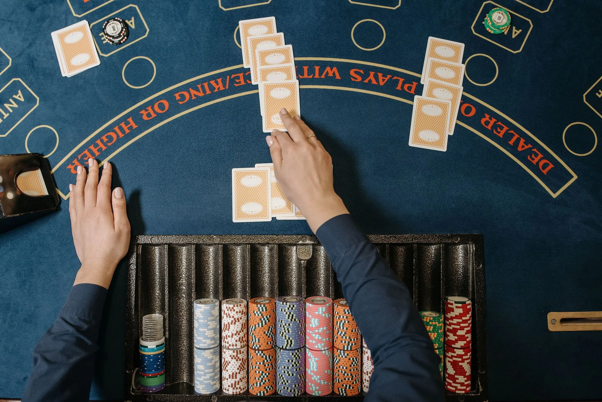A croupier's hands dealing cards across a blue baize casino table, with stacked chips in the tray below