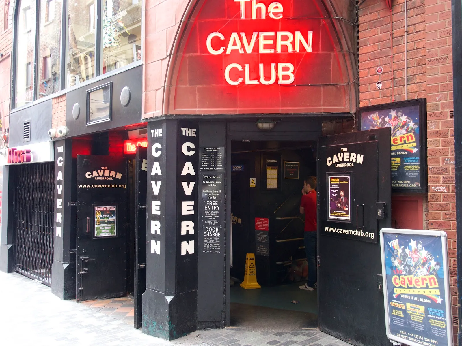 The Cavern Club's red neon sign above its Mathew Street entrance, with band posters and its own opening-times board beside the doorway