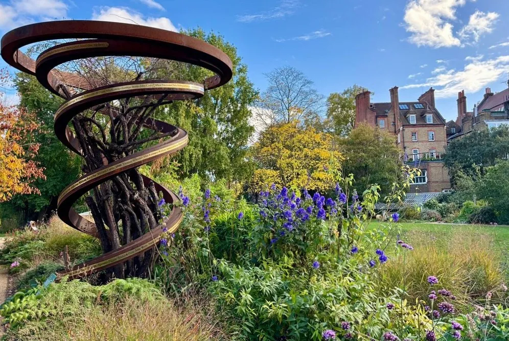 A spiral of rusted steel bands engraved with names, wound around a small tree at the Chelsea Physic Garden, with autumn borders of purple monkshood and verbena in front and a red-brick house beyond the wall