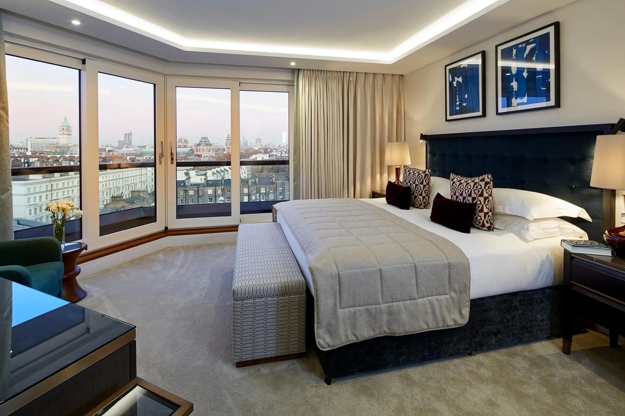 A bedroom at Cheval Gloucester Park, with a bow window looking out over London rooftops, a dark tufted headboard and a green armchair in the corner