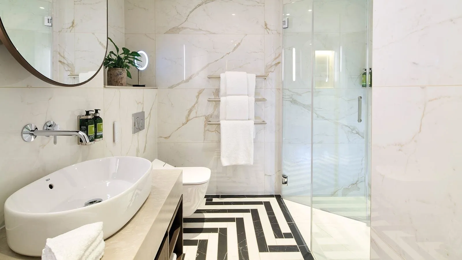 An apartment bathroom at Cheval Phoenix House in white marble-effect tiles, with a black and white herringbone floor, an oval basin, a walk-in shower behind glass and a heated towel rail