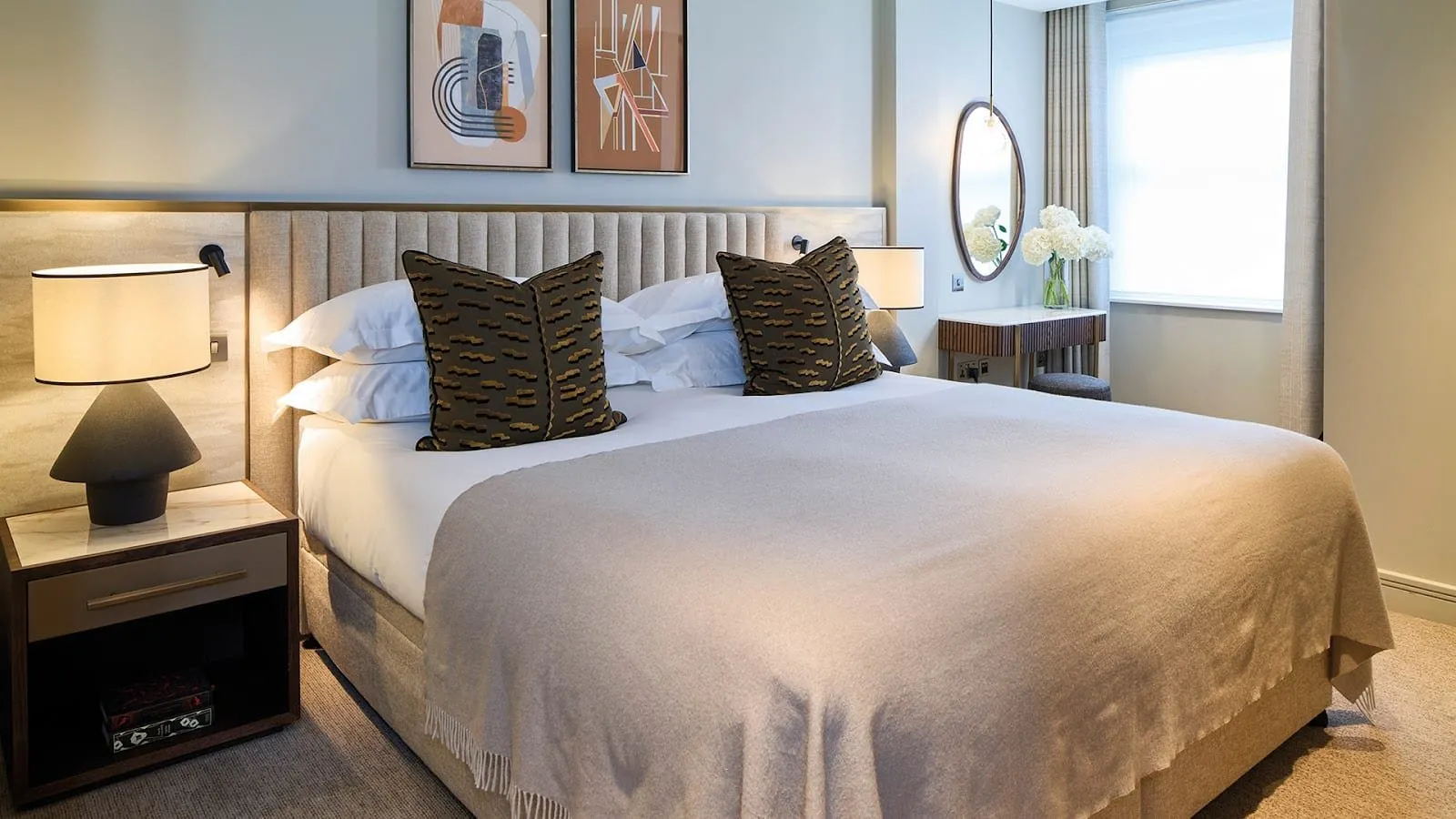 A bedroom at Cheval Phoenix House: a king bed with a channelled linen headboard, a cream wool throw, patterned brown cushions, two abstract prints, and a dressing table by the window