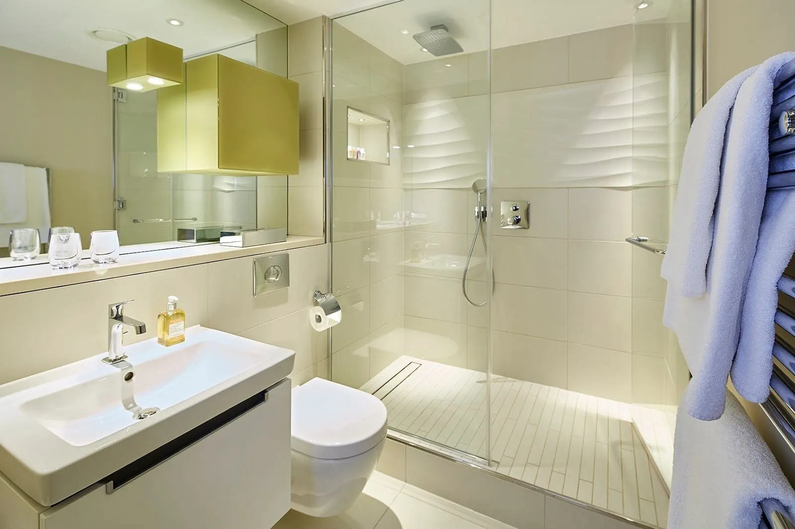 A bathroom at Cheval Three Quays with a walk-in rain shower behind glass, cream wave-textured tiles, a wall-hung toilet and a basin on a white vanity