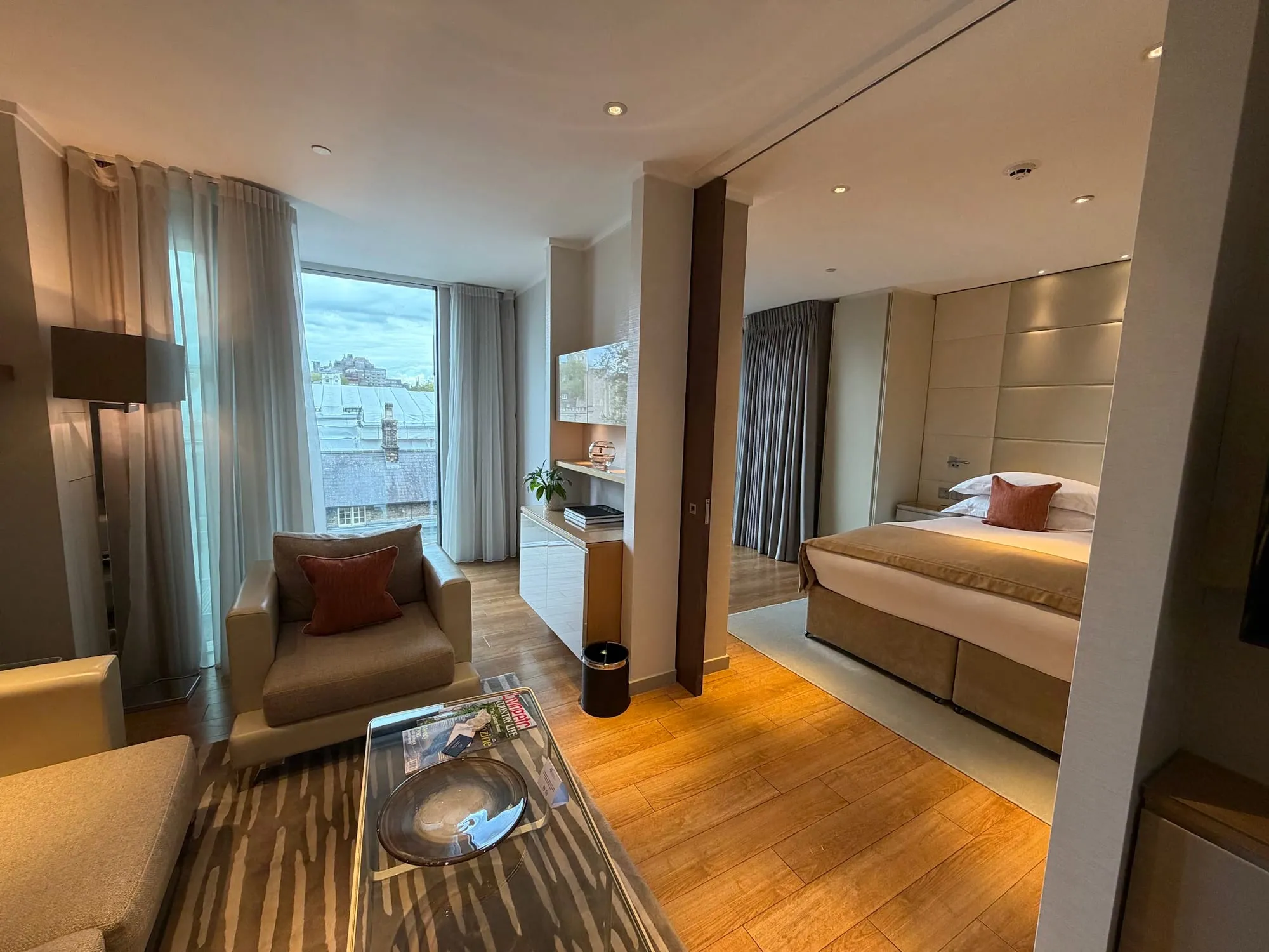 The living area of an apartment at Cheval Three Quays, with an armchair and coffee table in the foreground and the bedroom visible through an open partition beyond