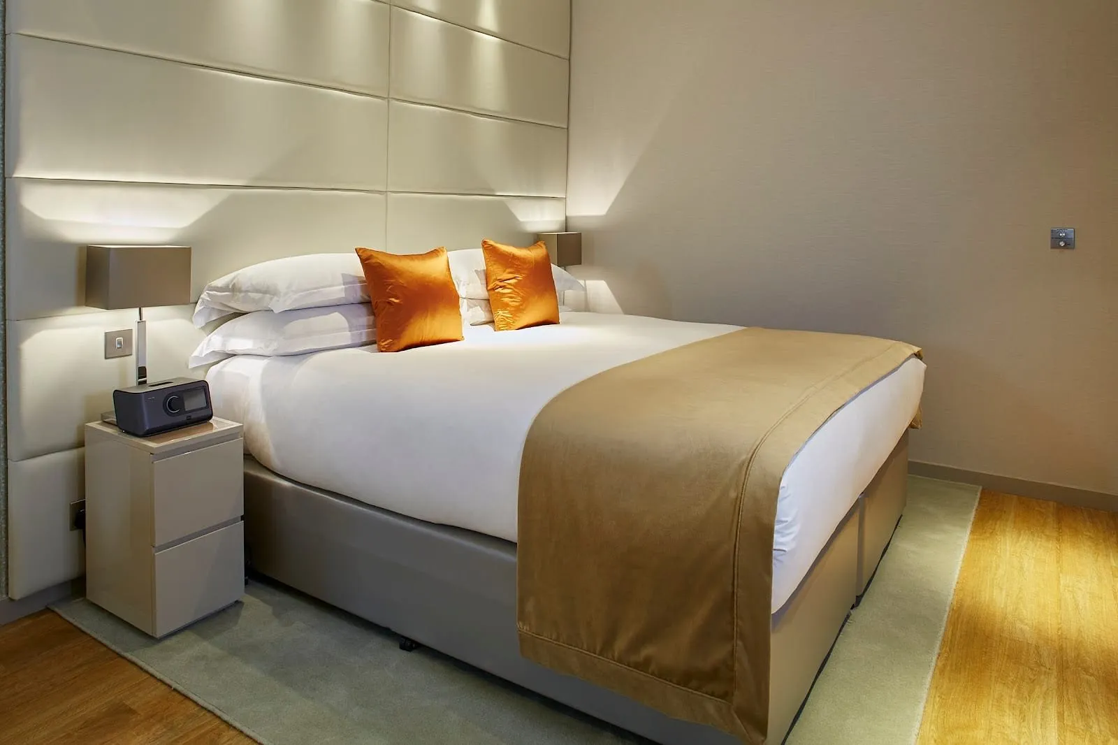 An apartment bedroom at Cheval Three Quays with a double bed against a padded cream leather headboard wall, orange silk cushions, a gold throw and oak floors
