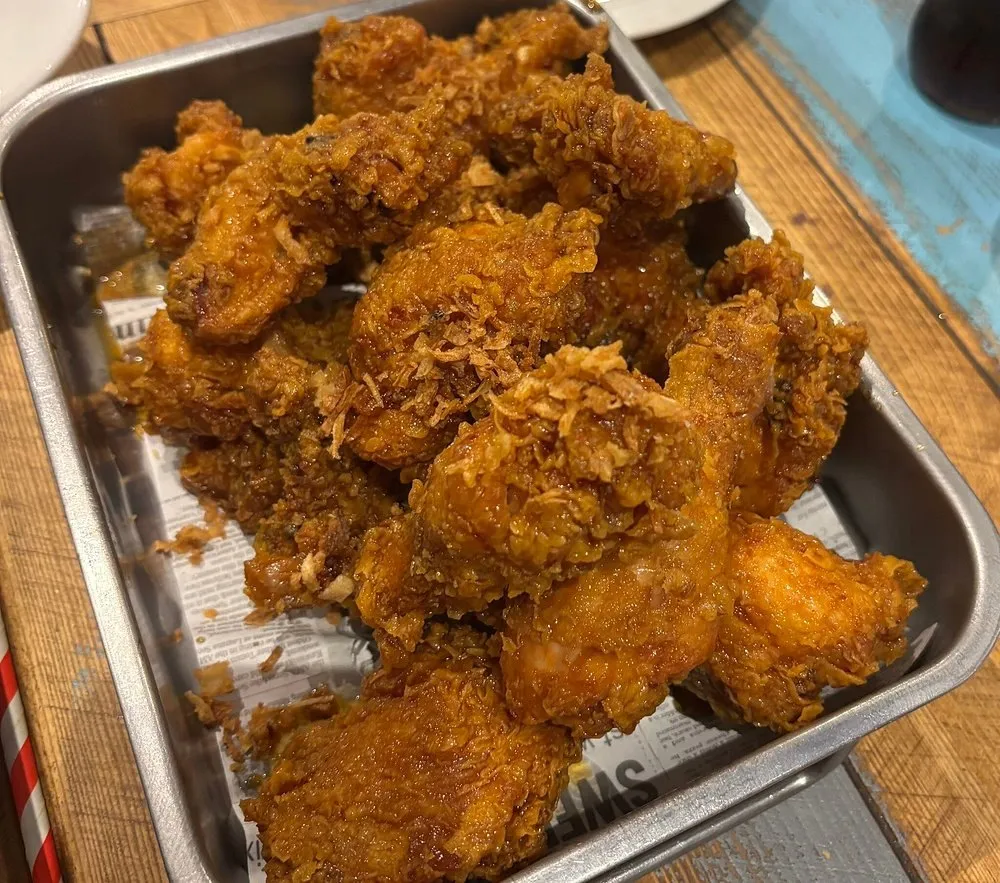 A metal tray of Korean fried chicken wings glazed and topped with crisp fried shallots, on newspaper