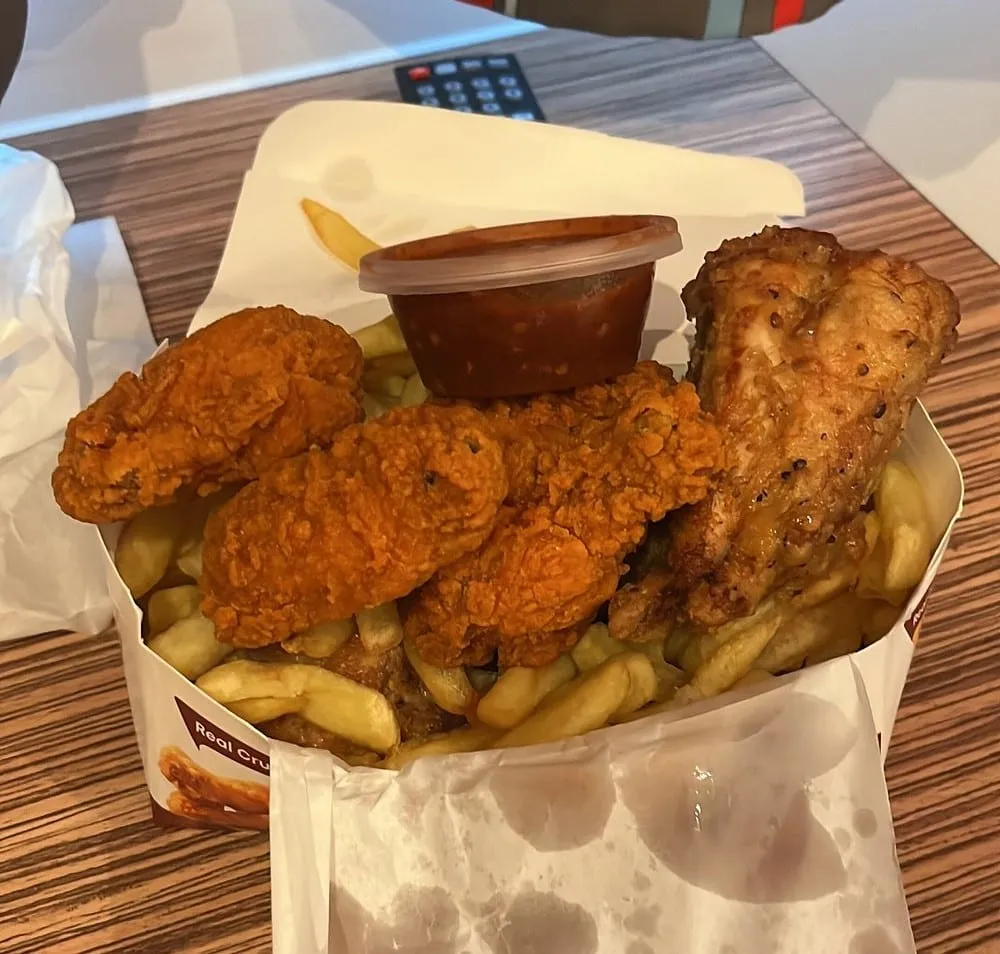 Fried chicken pieces, some crumbed red and some plain, piled over chips in a takeaway box with a pot of red sauce