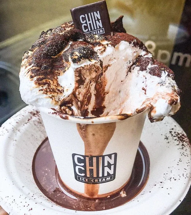 A paper cup of hot chocolate topped with a fist-sized marshmallow, blowtorched until charred and slumping over the rim, with a branded chocolate tag pushed into it
