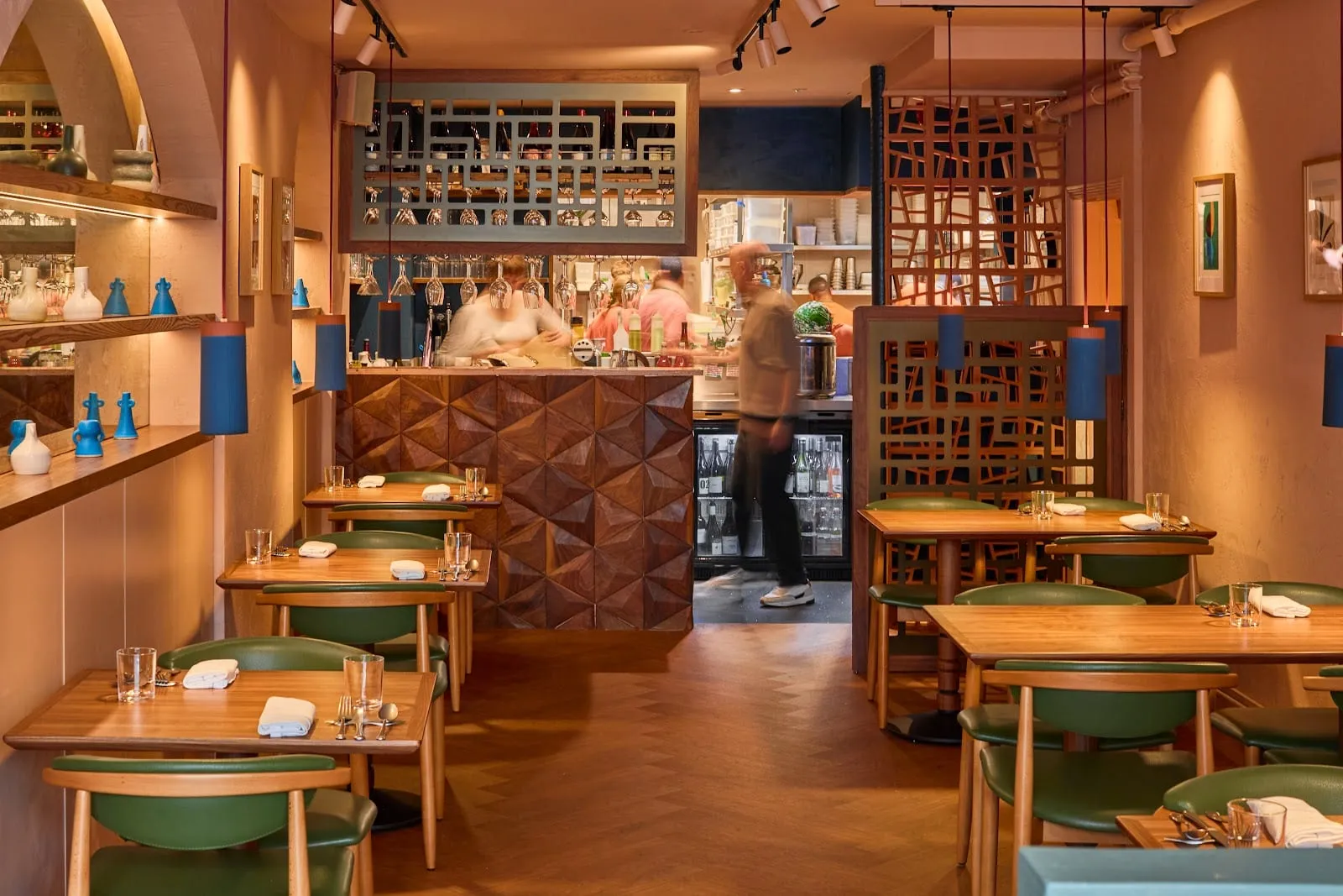 The dining room at Chishuru: green leather chairs at pale timber tables, a faceted wooden bar front, and glasses hanging above the open pass