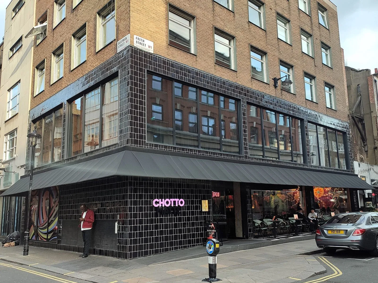 The corner of Frith Street and Bateman Street in Soho, with a black-tiled restaurant front, a pink neon Chotto Matte sign and diners at pavement tables under a grey awning