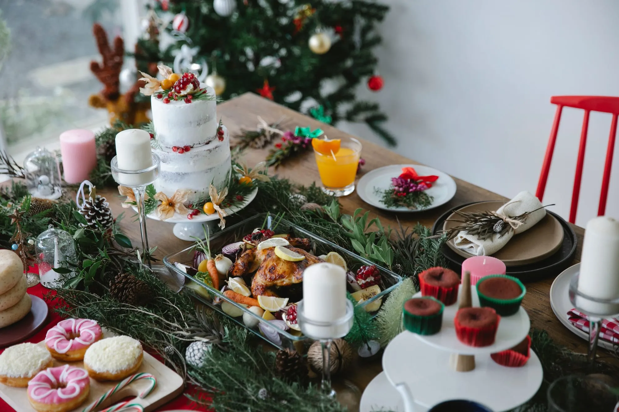 A Christmas table laid with a roast bird in a glass dish, a tiered iced cake, candles, cupcakes and pine foliage, with a decorated tree behind