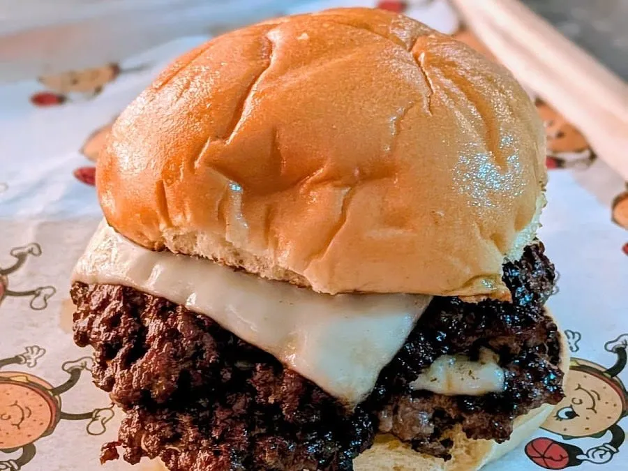 A Chuck's smash burger on paper printed with cartoon burgers: two heavily charred, crumbly-edged patties under a slice of melted white cheese in a shiny potato bun
